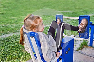 A little sporty girl in a sports uniform is engaged in the exercise machines on the playground for fitness on the street