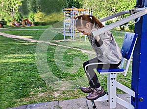 A little sporty girl in a sports uniform is engaged in the exercise machines on the playground for fitness on the street