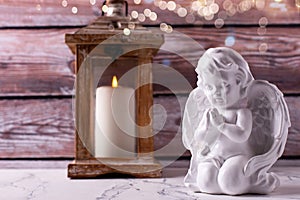 Little statue of white angle and lantern with burning candle  against brown textured wall.