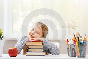 Little student girl sitting at the table. Tired schoolgirl with a pile of books. Education. Back to school. In the background is