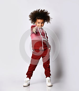 Little stylish African American girl in sportswear reaches out to you while standing against a gray background