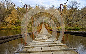 Little suspension pedestrian bridge over the river in the autumn forest. Outdoor hanging bridge made from wood and metal