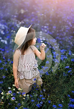 Little sweet cute girl in a hat with a flower in her hand looks away thoughtfully, standing in the middle of a meadow with bright