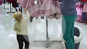 A little sweet girl with her mother chooses children`s clothes in a boutique. Mom and child choose clothes in the store