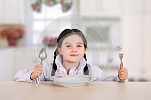 little teenage girl sitting at the dining table in the kitchen. Before eating with a plate, spoon, fork