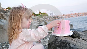 Little three year old girl eats cake on beach birthday pink dessert with three candles lick finger sit on stone pink bow