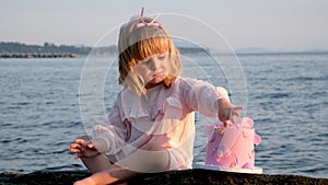 Little three year old girl eats cake on beach birthday pink dessert with three candles lick finger sit on stone pink bow