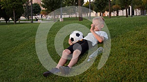 A little tired boy is lying on the green grass in a city park with a soccer ball