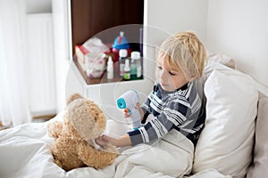 Little toddler blond child, playing with teddy bear in bed, while being sick, checking his temperature