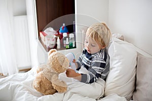 Little toddler blond child, playing with teddy bear in bed, while being sick, checking his temperature