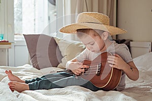 Little toddler boy in hat playing ukulele guitar at home, rustic style. Lifestyle concept