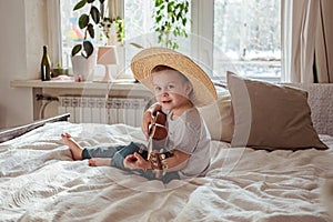 Little toddler boy in hat playing ukulele guitar at home, rustic style. Lifestyle concept