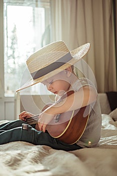 Little toddler boy in hat playing ukulele guitar at home, rustic style. Lifestyle concept