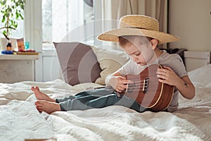 Little toddler boy in hat playing ukulele guitar at home, rustic style. Lifestyle concept