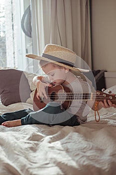 Little toddler boy in hat playing ukulele guitar at home, rustic style. Lifestyle concept