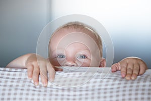 Little toddler boy playing on the bed. Cute kid smiling and hiding under cover. Palyful and mischievous eyes. Hide-and-seek.