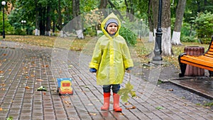 Little toddler boy in raincoat and rubber boots got upset because of bad weather and heavy rain in park