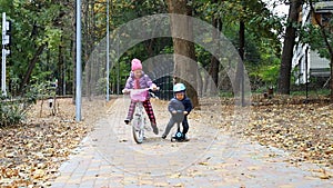 Little toddler boy riding scooter balance bike by asphalt walkway together with mother walking near path at city park at