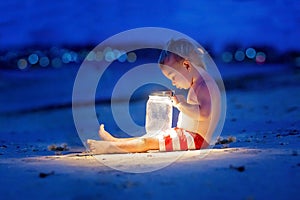 Little toddler boy, sitting on the beach after sunset on a moonlight with a lantern, enjoying the quiteness of the beach