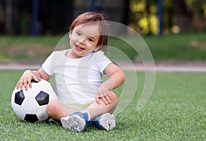 Little toddler boy sitting with legs crossed on football field in summer day with soccer ball. Happy active child