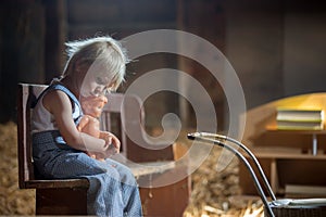 Little toddler boy, sitting on old vintage bench, holding doll  in attic