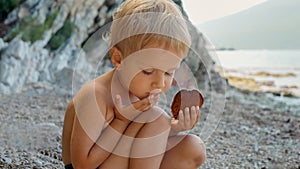 Little toddler boy sitting on the sea beach and eating pancakes