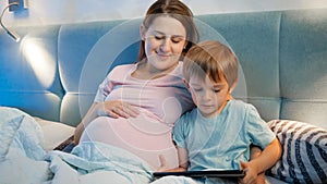 Little toddler boy using tablet computer lying with pregnant mother in bed at night