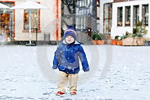 Little toddler boy walking through the snowy city during snowfall. Cute happy child in winter clothes having fun