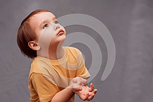 Little toddler boy in yellow shirt standing and looking up asking for something. The kid is holding hands raised to chest like he