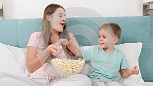 Little toddler boy with young mother in pajamas lying in bed on weekend and eating popcorn from big bowl. Concept of