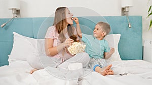 Little toddler boy with young mother in pajamas lying in bed on weekend and eating popcorn from big bowl. Concept of