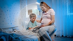 Little toddler boy with young pregnant mother playing games and watching cartoons on tablet computer in bed before going