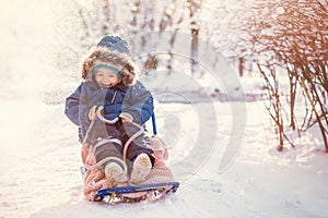 Happy toddler kid sits on sled under snowfall and laughs hiding from snow. Toned image. Place for copy text