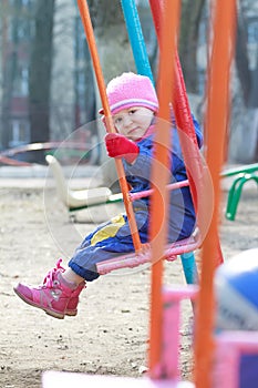 Little toddler girl in dark blue warm overall is sitting on playgrounds swing