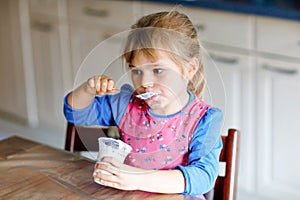 Little toddler girl eating yogurt for breakfast. Cute healthy baby sitting in the kitchen or at nursery and having meal.