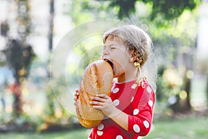 Little toddler girl holding big loaf of bread. Funny happy child biting and eating healthy bread, outdoors. Hungry kid.