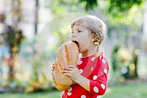 Little toddler girl holding big loaf of bread. Funny happy child biting and eating healthy bread, outdoors. Hungry kid. photo