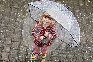 Little toddler girl playing with big umbrella on rainy day. Happy positive child running through rain, puddles
