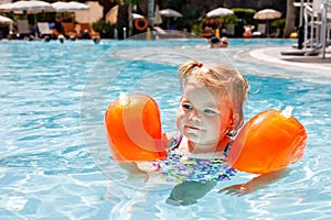 Little toddler girl with protective swimmies playing in outdoor swimming pool by sunset. Baby Child learning to swim in