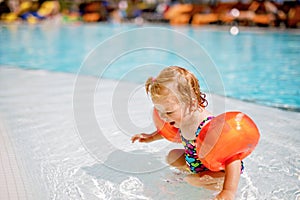 Little toddler girl with protective swimmies playing in outdoor swimming pool by sunset. Baby Child learning to swim in