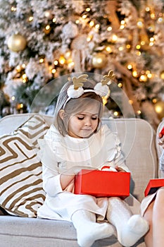 A little toddler girl sits on a sofa near a decorated Christmas tree and opens presents. Christmas Holidays