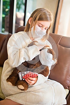 Little toddler girl sitting in medical mask with teddy bear feeling sick with flu and fever