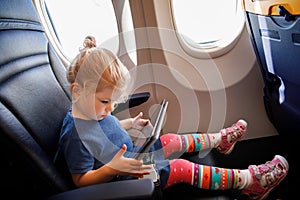 Little toddler girl traveling by plane. Small happy child sitting by aircraft window and using a digital tablet during