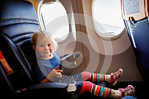 Little toddler girl traveling by plane. Small happy child sitting by aircraft window and using a digital tablet during