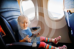 Little toddler girl traveling by plane. Small happy child sitting by aircraft window and using a digital tablet during