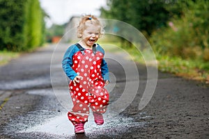 Little toddler girl wearing rain boots and trousers and walking during sleet, rain on cold day. Baby child having fun