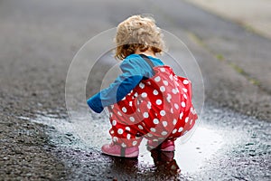 Little toddler girl wearing rain boots and trousers and walking during sleet, rain on cold day. Baby child having fun