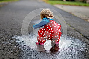 Little toddler girl wearing rain boots and trousers and walking during sleet, rain on cold day. Baby child having fun