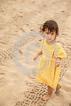 Little toddler girl in yellow dress walking on sandy beach at summer time, childhood memories