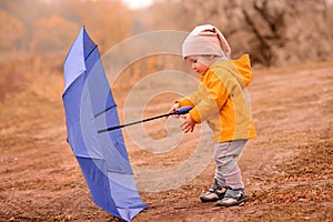 Little toddler girl in yellow raincoat playing with big blue umbrella outdoors in autumn day